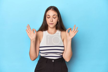 Photo for Woman in a striped top posing on a blue studio backdrop relaxes after hard working day, she is performing yoga. - Royalty Free Image