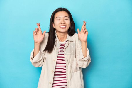 Téléchargez les photos : Femme asiatique en chemise à couches et t-shirt rayé, croisant les doigts pour avoir de la chance - en image libre de droit