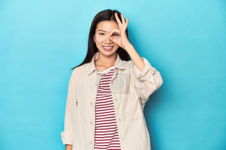 Foto de Mujer asiática con camisa en capas y camiseta a rayas, emocionada manteniendo el gesto bien en el ojo. - Imagen libre de derechos