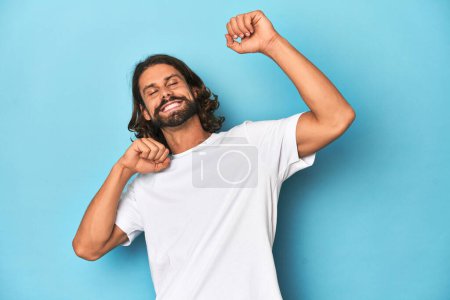 Foto de Hombre barbudo con camisa blanca, fondo azul celebrando un día especial, salta y levanta los brazos con energía. - Imagen libre de derechos