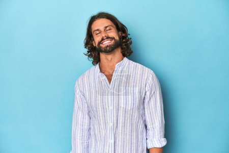 Photo for Man with beard in blue striped shirt, blue studio laughs and closes eyes, feels relaxed and happy. - Royalty Free Image