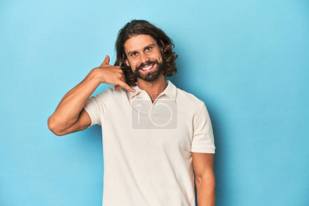 Téléchargez les photos : Homme aux cheveux longs dans un polo blanc, studio bleu montrant un geste d'appel téléphonique mobile avec les doigts. - en image libre de droit
