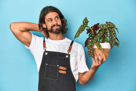 Téléchargez les photos : Jardinier aux cheveux longs avec barbe tenant une plante touchant l'arrière de la tête, pensant et faisant un choix. - en image libre de droit