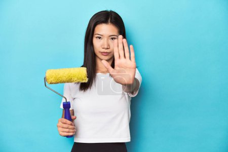 Téléchargez les photos : Jeune femme asiatique avec rouleau de peinture, concept de bricolage, debout avec la main tendue montrant le signe d'arrêt, vous empêchant. - en image libre de droit