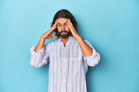 Photo for Man with beard in blue striped shirt, blue studio touching temples and having headache. - Royalty Free Image