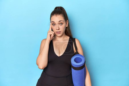 Photo for Young sporty Caucasian woman with yoga mat in blue studio Young sporty Caucasian woman with yoga mat in blue studiopointing temple with finger, thinking, focused on a task. - Royalty Free Image