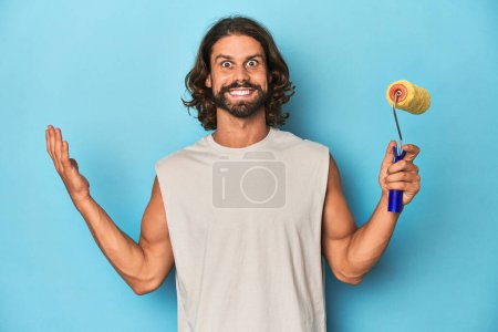 Photo for Bearded man painting with a yellow roller receiving a pleasant surprise, excited and raising hands. - Royalty Free Image