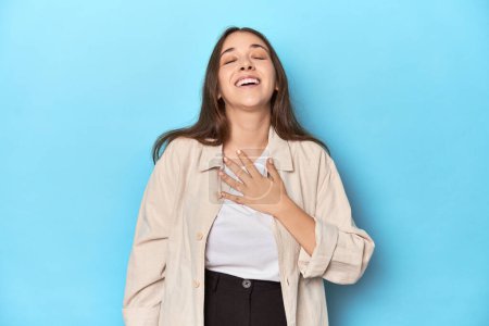 Foto de Mujer joven con estilo en una camisa sobre un fondo azul se ríe en voz alta manteniendo la mano en el pecho. - Imagen libre de derechos