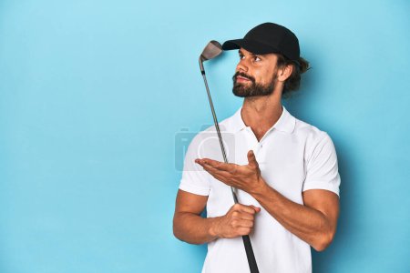 Photo for Bearded, long-haired golfer holding a golf club in a blue studio. - Royalty Free Image