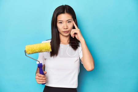 Téléchargez les photos : Jeune femme asiatique avec rouleau de peinture, concept de bricolage, pointant le temple avec le doigt, pensant, concentré sur une tâche. - en image libre de droit
