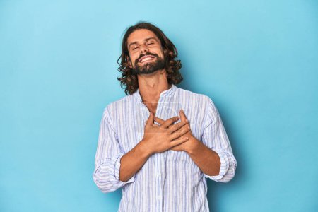 Foto de Hombre con barba en camisa de rayas azules, estudio azul riendo manteniendo las manos en el corazón, concepto de felicidad. - Imagen libre de derechos