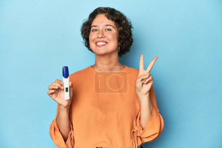 Photo for Young woman holding pregnancy test, studio background showing number two with fingers. - Royalty Free Image
