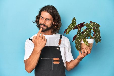 Foto de Jardinero de pelo largo con barba sosteniendo una planta apuntándote con el dedo como si invitaras a acercarte. - Imagen libre de derechos