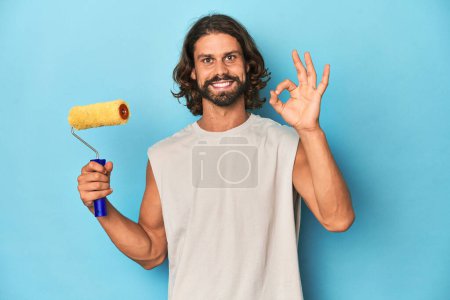 Photo for Bearded man painting with a yellow roller cheerful and confident showing ok gesture. - Royalty Free Image