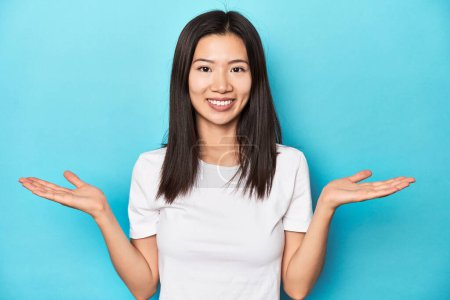 Téléchargez les photos : Jeune femme asiatique en t-shirt blanc, prise de vue en studio, fait échelle avec les bras, se sent heureux et confiant. - en image libre de droit