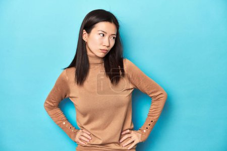 Photo for Young Asian woman in brown turtleneck, confused, feels doubtful and unsure. - Royalty Free Image