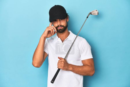 Foto de Golfista de pelo largo con club y sombrero apuntando templo con el dedo, pensando, centrado en una tarea. - Imagen libre de derechos