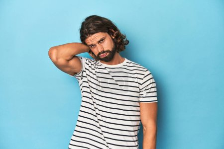 Photo for Bearded man in a striped shirt, blue backdrop suffering neck pain due to sedentary lifestyle. - Royalty Free Image