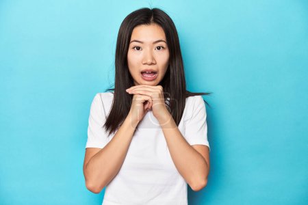 Foto de Mujer asiática joven en camiseta blanca, plano de estudio, rezando por suerte, asombrada y con la boca abierta mirando al frente. - Imagen libre de derechos