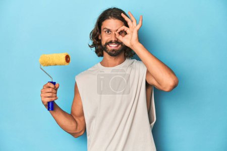 Photo for Bearded man painting with a yellow roller excited keeping ok gesture on eye. - Royalty Free Image