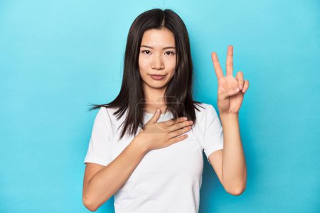 Photo for Young Asian woman in white t-shirt, studio shot, taking an oath, putting hand on chest. - Royalty Free Image