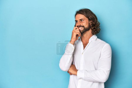 Téléchargez les photos : Homme barbu dans une chemise blanche, fond bleu détendu penser à quelque chose qui regarde un espace de copie. - en image libre de droit