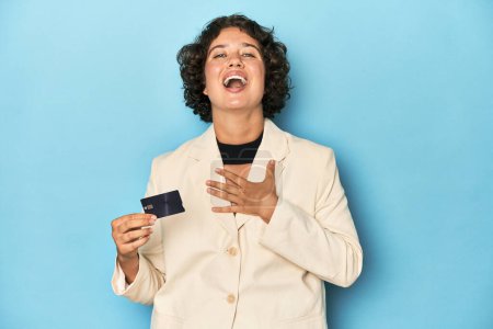 Photo for Young woman with credit card, elegant white blazer laughs out loudly keeping hand on chest. - Royalty Free Image