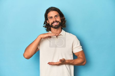 Foto de Hombre de pelo largo en un polo blanco, estudio azul sosteniendo algo con ambas manos, presentación del producto. - Imagen libre de derechos