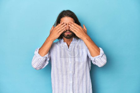 Photo for Man with beard in blue striped shirt, blue studio afraid covering eyes with hands. - Royalty Free Image