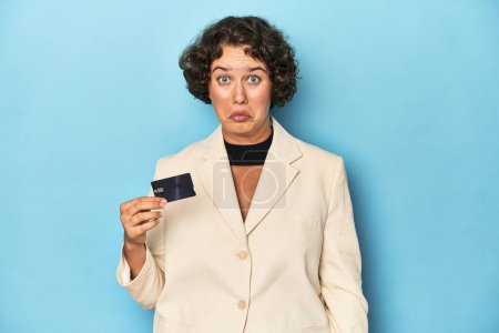 Photo for Young woman with credit card, elegant white blazer shrugs shoulders and open eyes confused. - Royalty Free Image