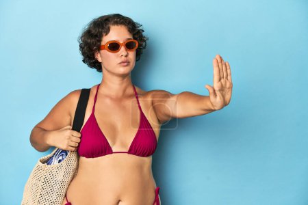 Foto de Mujer joven en bikini con bolsa de playa de pie con la mano extendida mostrando señal de stop, lo que le impide. - Imagen libre de derechos