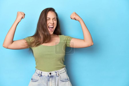 Photo for Young Caucasian woman in a green top on a blue backdrop raising fist after a victory, winner concept. - Royalty Free Image