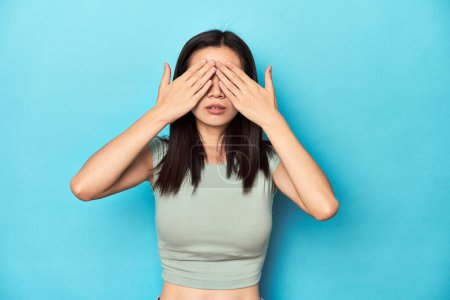 Photo for Asian woman in summer green top, studio backdrop, afraid covering eyes with hands. - Royalty Free Image