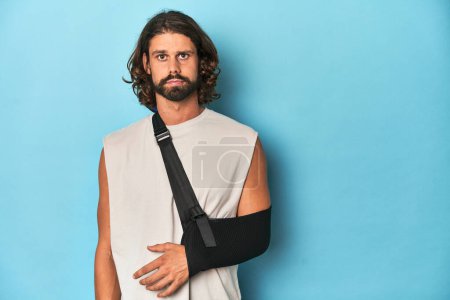 Photo for Man with a beard, long hair and arm in sling looking pained in blue studio. - Royalty Free Image