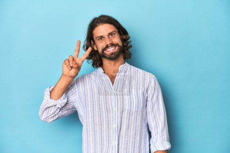 Photo for Man with beard in blue striped shirt, blue studio joyful and carefree showing a peace symbol with fingers. - Royalty Free Image