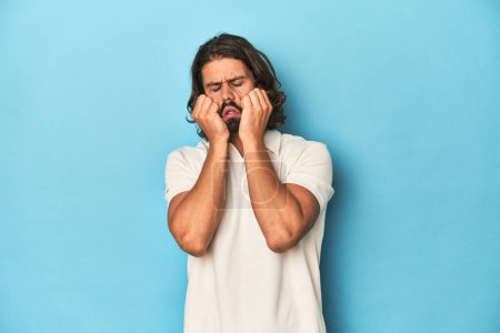 Photo for Long-haired man in a white polo, blue studio crying, unhappy with something, agony and confusion concept. - Royalty Free Image