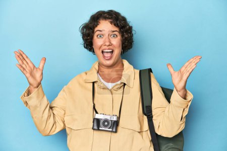 Téléchargez les photos : Jeune voyageuse avec caméra vintage recevant une agréable surprise, excitée et levant les mains. - en image libre de droit