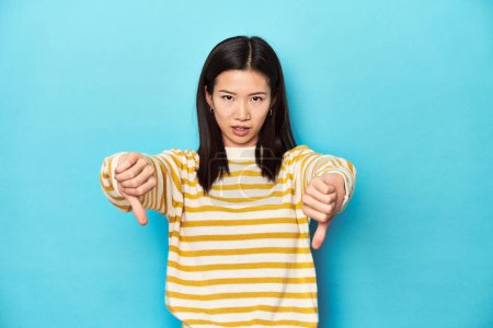 Photo for Asian woman in striped yellow sweater, showing thumb down and expressing dislike. - Royalty Free Image