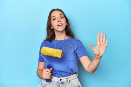Photo for Young woman with yellow paint roller on a blue background smiling cheerful showing number five with fingers. - Royalty Free Image
