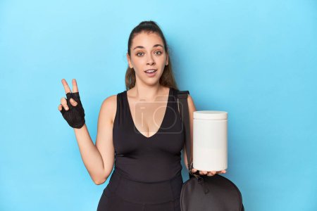 Téléchargez les photos : Jeune femme tenant une bouteille de protéines dans un cadre sportif montrant le numéro deux avec les doigts. - en image libre de droit