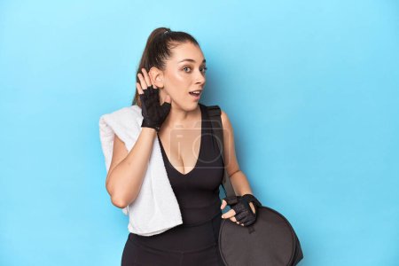 Téléchargez les photos : Jeune femme prête pour la gym avec des éléments essentiels sur fond bleu en essayant d'écouter un potins. - en image libre de droit