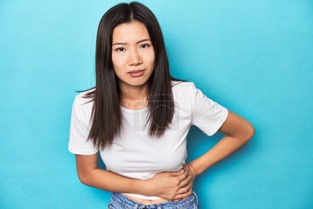 Foto de Mujer asiática joven en camiseta blanca, toma de estudio, tiene un dolor de hígado, dolor de estómago. - Imagen libre de derechos