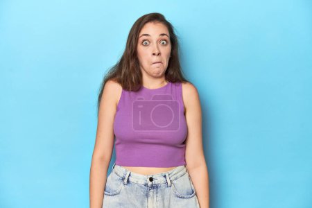 Foto de La mujer a la moda joven en la parte superior púrpura sobre el fondo azul encoge los hombros y abre los ojos confundido. - Imagen libre de derechos