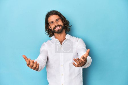 Photo for Bearded man in a white shirt, blue backdrop showing a welcome expression. - Royalty Free Image