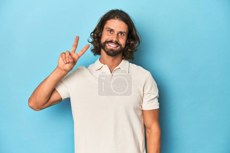 Téléchargez les photos : Homme aux cheveux longs dans un polo blanc, studio bleu joyeux et insouciant montrant un symbole de paix avec les doigts. - en image libre de droit