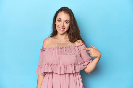 Téléchargez les photos : Jeune femme portant une robe rose sur fond bleu studio personne pointant à la main vers un espace de copie chemise, fière et confiante - en image libre de droit