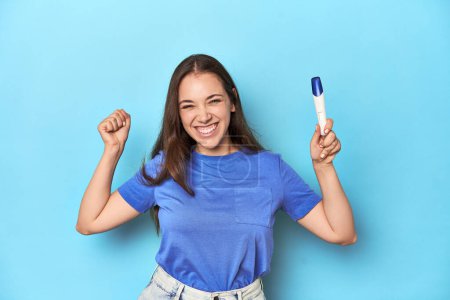 Foto de Mujer emocionada sosteniendo una prueba de embarazo positiva en estudio azul. - Imagen libre de derechos