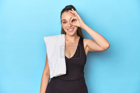 Téléchargez les photos : Fit jeune femme avec une serviette sur son épaule, studio tourné excité en gardant ok geste sur l'œil. - en image libre de droit