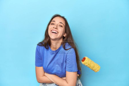 Foto de Mujer joven con rodillo de pintura amarillo sobre un fondo azul riendo y divirtiéndose. - Imagen libre de derechos