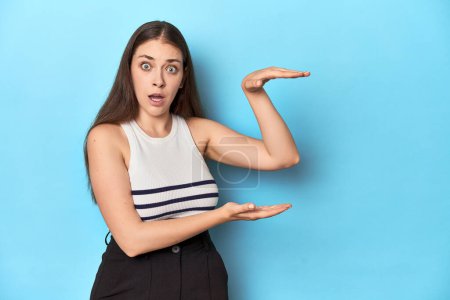Photo for Woman in a striped top posing on a blue studio backdrop shocked and amazed holding a copy space between hands. - Royalty Free Image